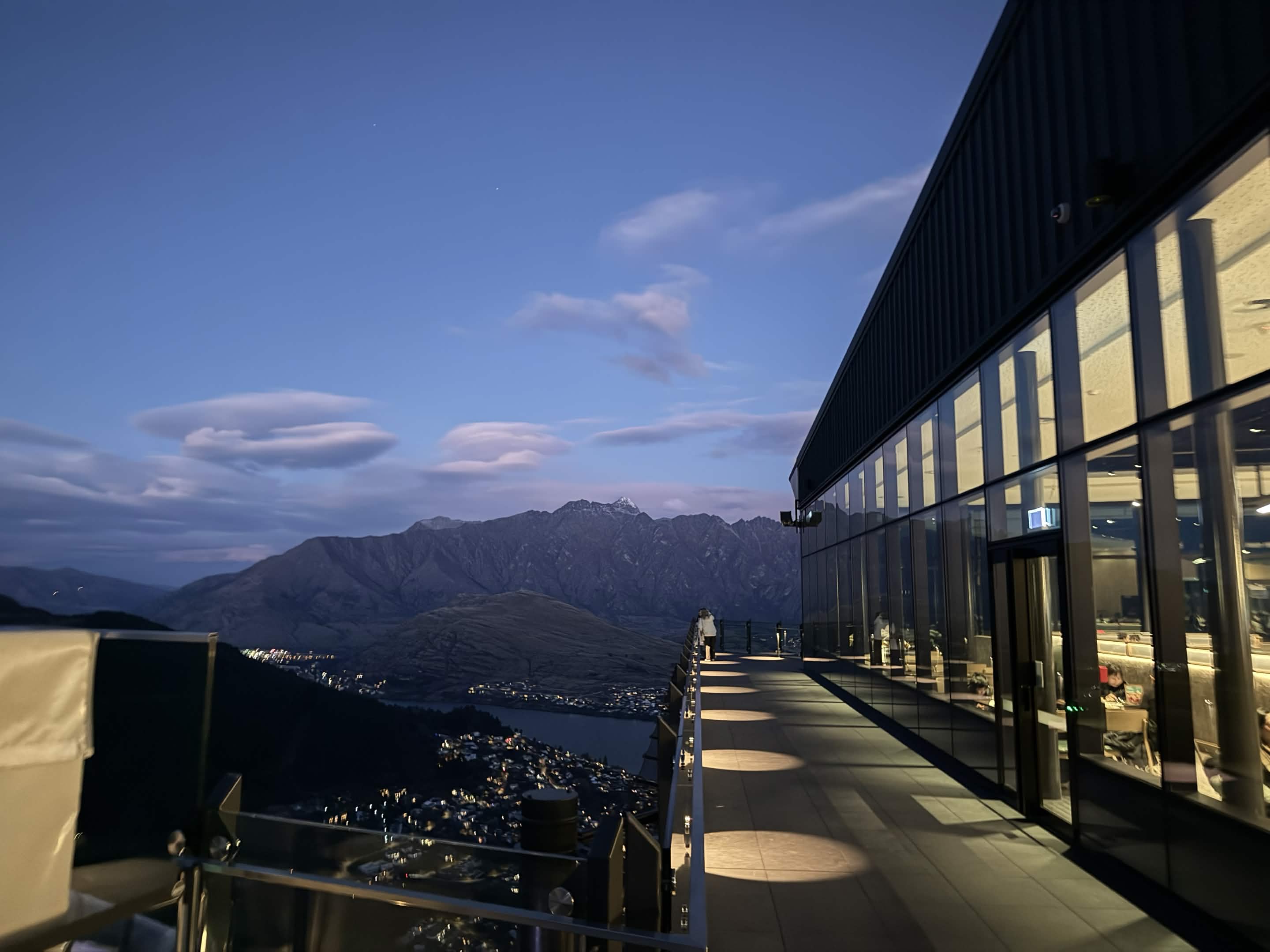 Blue-hour view from the Skyline summit terrace, the glass-walled restaurant lit up on the right and Queenstown's lights below with The Remarkables in the distance